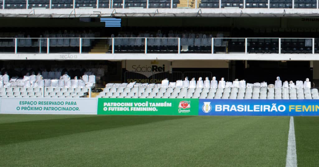 Foto do campo de futebol do estádio do Santos FC. Na imagem se vê uma placa de Guaraná Antarctica com os dizeres: 'Espaço reservado para o próximo patrocinador. Patrocine você também o futebol feminino.".