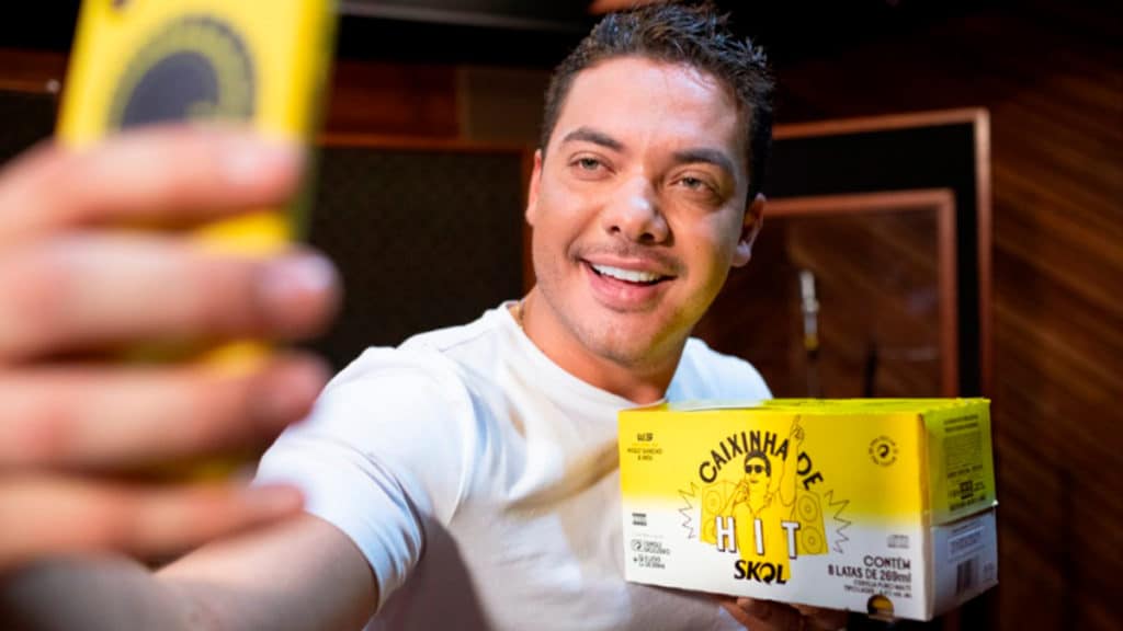 Um homem sorridente tira uma selfie enquanto segura uma caixa de cerveja Skol, com um ambiente de estúdio ao fundo.