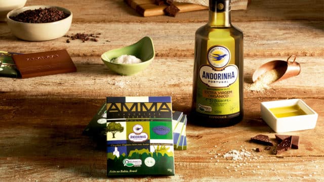 Uma mesa com os ingredientes do chocolate espalhados nela, no centro o chocolate com azeite da AMMA e ao lado uma garrafa de azeite Andorinha Extra Virgem.