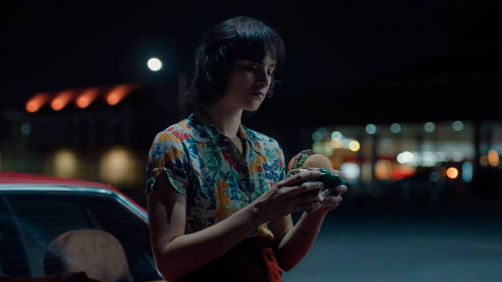 burger-king-whopper-de-plantas Foto da campanha de Burger King para o Whopper de Plantas. A foto apresenta uma mulher encostada em seu carro, durante a noite, prestes a comer o Whopper de Plantas.