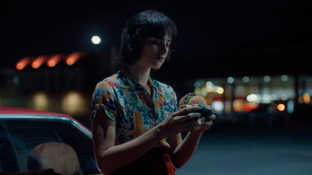 Foto da campanha de Burger King para o Whopper de Plantas. A foto apresenta uma mulher encostada em seu carro, durante a noite, prestes a comer o Whopper de Plantas.