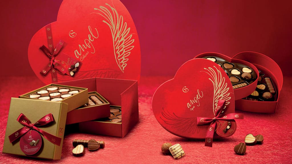 Foto de divulgação da linha de Dia das Mães da Cacau Show. A foto apresenta um fundo e uma mesa na cor vermelha, com as Caixas Angel Coleção, Cupido e Porta-Joias, e alguns bombons de chocolate branco e preto espalhados pela mesa.