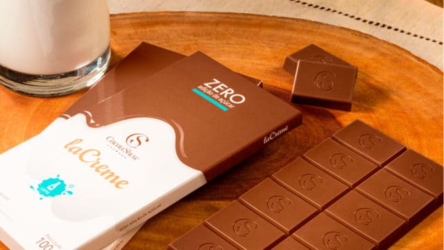 Tabletes de laCreme ao Leite Zero Adição de Açúcar da Cacau Show em uma mesa ao lado de um copo de leite.