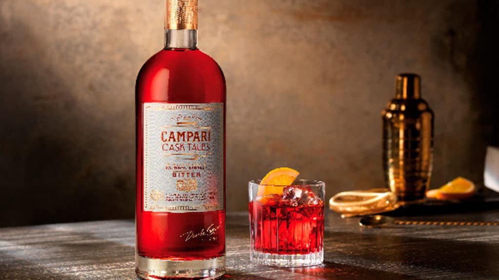 Uma garrafa de Campari Cask Tales, ao lado um copo com a bebida, com gelo e uma fatia de laranja. Ao fundo uma coqueteleira com fatias de laranjas.