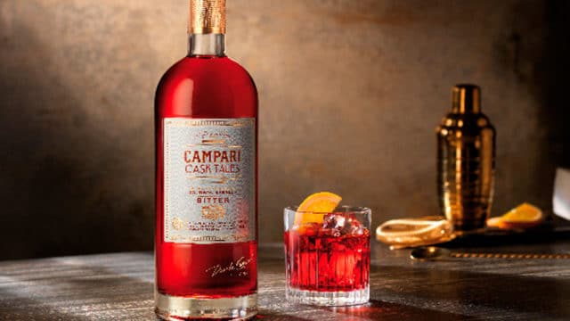 Uma garrafa de Campari Cask Tales, ao lado um copo com a bebida, com gelo e uma fatia de laranja. Ao fundo uma coqueteleira com fatias de laranjas.