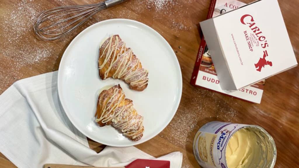 Foto de divulgação da linha de Dia das Mães da Carlo's Bakery com Leite Moça. Na foto, uma mesa com duas Lobster Tails Feito Com Moça, ao lado de um livro, uma caixa da loja, uma lata de Leite Moça e utensílios de confeitaria.