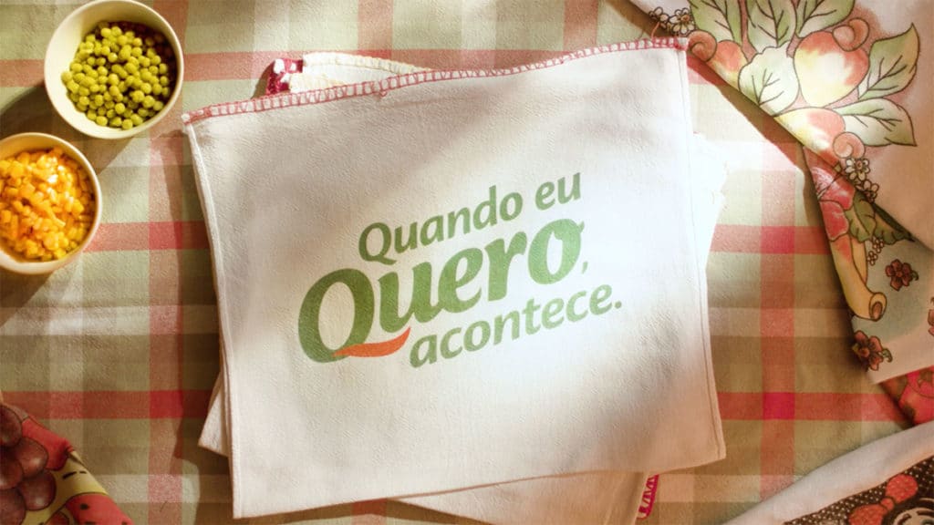 Banner de divulgação da nova campanha da Quero, que exalta a força de vontade das consumidoras. Foto com a frase "Quando eu Quero acontece" bordado em um pano de prato em uma mesa com temperos.