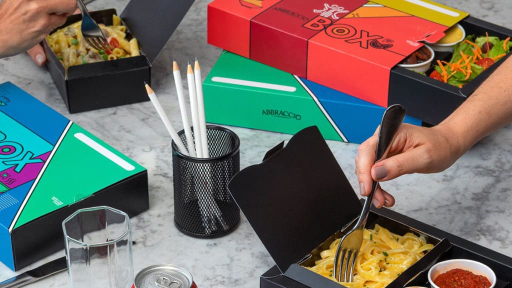 A foto apresenta uma mesa com vários Abbraccio Box espalhados e duas mãos dando uma garfada em dois boxes abertos.