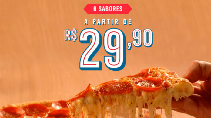 Foto de divulgação da promoção de pizza por R$ 29,90 da Domino's. A foto apresenta uma pessoa pegando um pegado de pizza de Pepperoni, e acima está escrito