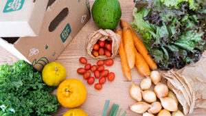 Foto de divulgação do mercado online da Liv Up. A foto apresenta uma mesa de madeira cheia de legumes e verduras espalhas, junto a uma caixa de papelão da Liv Up.