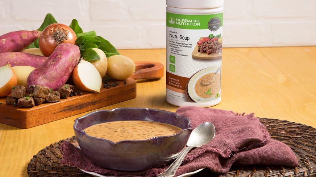 nutri-soup-carne-batata A foto apresenta uma mesa montada com uma tábua de batatas, um bowl com sopa, um jogo americano e uma colher, além de um pote de Nutri Soup Creme de Carne com Batata Doce