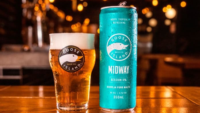 Goose Island Midway lata ao lado de um copo com cerveja, em um balcão de bar, ao fundo luzes com um ambiente desfocado.