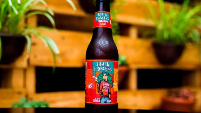 A foto apresenta a nova cerveja da Black Princess feita apenas por mulheres em cima de uma mesa de madeira, com várias plantas e paletes atrás.