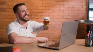 Foto de divulgação da ação em que Brahma procura um consumidor para tomar chopps de cerveja, a fotografia apresenta o humorista Maurício Meirelles tomando uma cerveja em frente ao notebook.