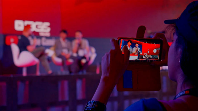 A foto apresenta uma menina tirando uma fotografia de um painel que aconteceu em edições passadas da Brasil Game Show, para representar uma das atrações da programação do evento.