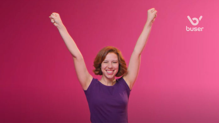 A foto apresenta um frame da campanha de posicionamento da Buser, em que mostra uma mulher sorrindo com os braços levantados, ao lado do logo da marca.