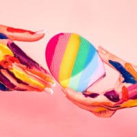 A foto da campanha para celebrar o orgulho LGBTQIAP+ da FOREO apresenta um fundo rosa, e duas mãos na frente com várias pinceladas coloridas, passando a YOUnique LUNA de uma para a outra.