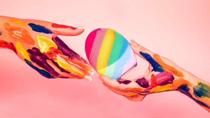 A foto da campanha para celebrar o orgulho LGBTQIAP+ da FOREO apresenta um fundo rosa, e duas mãos na frente com várias pinceladas coloridas, passando a YOUnique LUNA de uma para a outra.