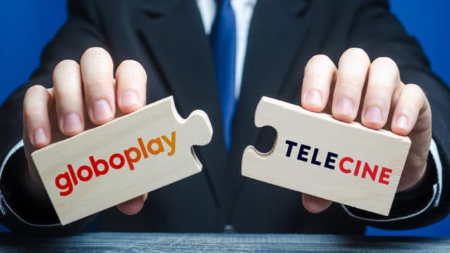 A foto apresenta um close na mão de um executivo segurando duas peças de um quebra-cabeça que se encaixam perfeitamente, em cada uma delas têm o logo do Globoplay e no outro do Telecine.