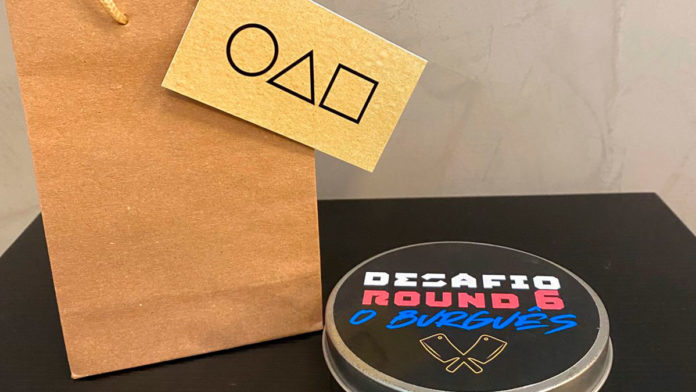 A foto apresenta uma sacola de papel com um cartão de visitas com um círculo, um triângulo e um quadrado, e um pote do desafio do biscoito de Round 6 com o nome e logo da hamburgueria O Burguês.