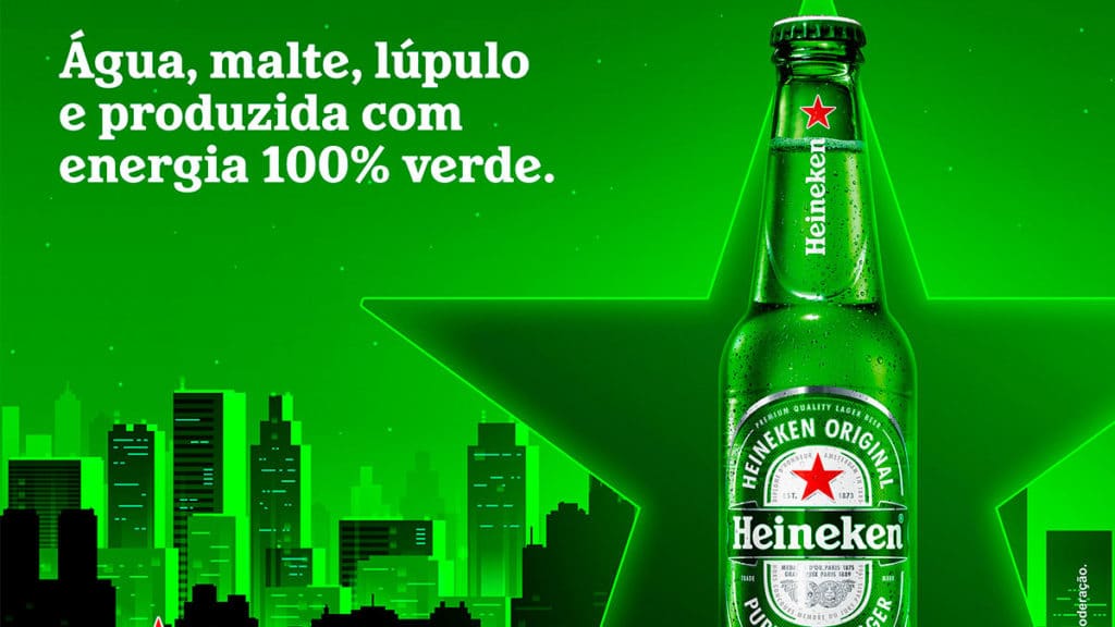 A foto apresenta um fundo com uma cidade desenhada e toda iluminada de verde com uma garrada de Heineken em frente sendo destacada por uma estrela e um texto no canto superior esquerdo. O texto diz "Água, malte, lúpulo e produzida 100% com energia verde".