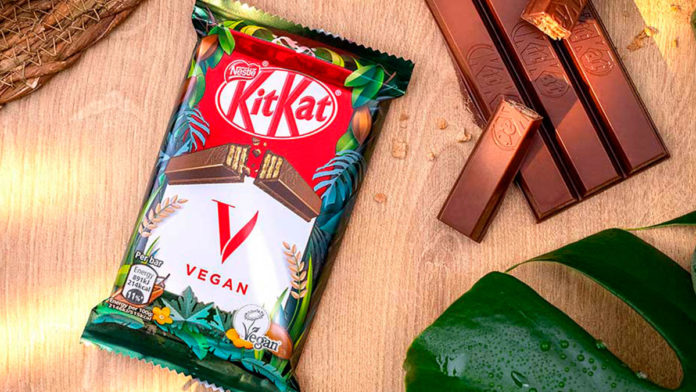 A foto apresenta uma embalagem do KitKat V em uma mesa de madeira, ao lado do chocolate vegano desembalado e algumas plantas ao redor.