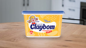 A foto apresenta uma Margarina sabor Manteiga Claybom 1kg em cima de uma mesa de madeira, em uma cozinha.