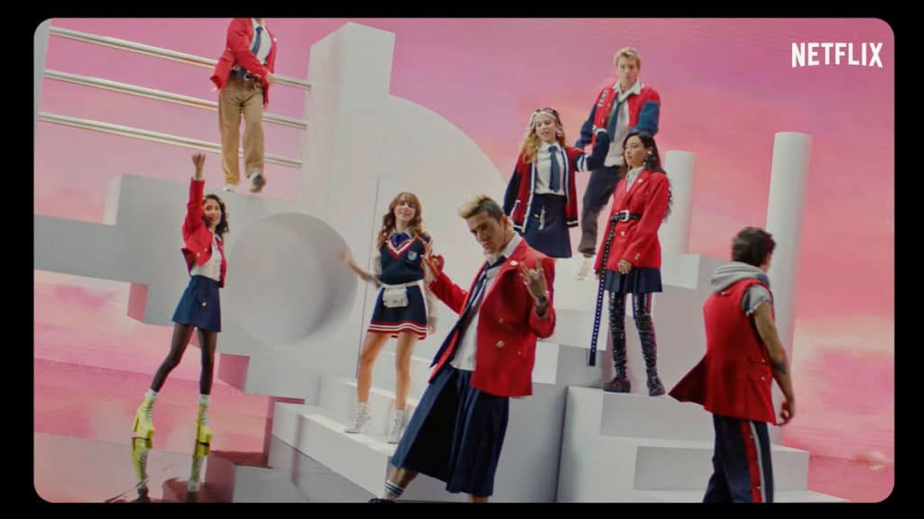 netflix-rebelde A foto apresenta o elenco de Rebelde Netflix cantando em pé em um cenário cheio de escadas brancas e fundo rosa.
