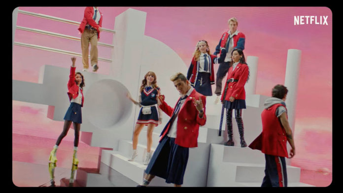 A foto apresenta o elenco de Rebelde Netflix cantando em pé em um cenário cheio de escadas brancas e fundo rosa.