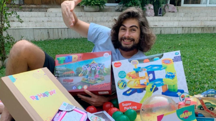 A foto apresenta o ator Rafa Vitti na grama deitado e sorrindo, com a mão levantada, junto com vários brinquedos do Grupo Ri Happy.
