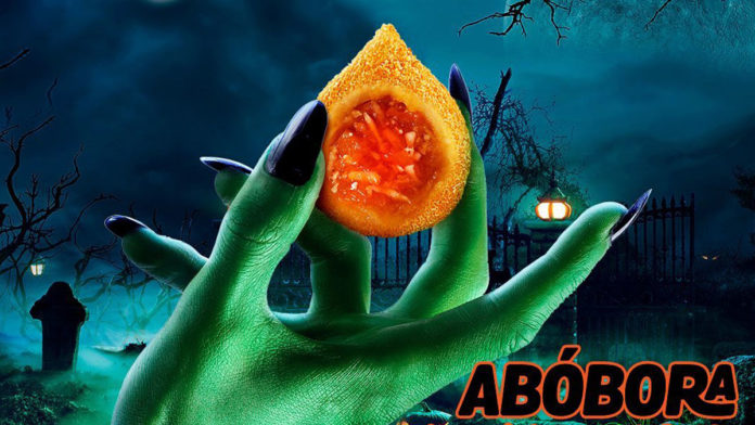 A foto apresenta uma mão verde segurando a Coxinha de Abóbora com Coco do Ragazzo, com um fundo de um cemitério a noite e a palavra abóbora ao lado.