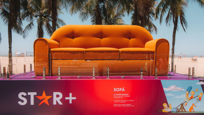 A foto apresenta a escultura do sofá de Os Simpsons que a Star+ colocou na Orla de Copacabana, em meio a vários coqueiros.