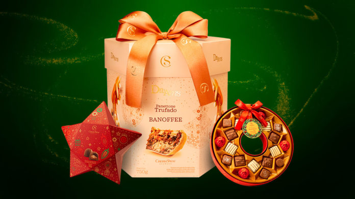 A foto apresenta o novo Panettone de Banoffee da Cacau Show entre duas caixas de bombons de chocolate, uma em formato de estrela e outra de guirlanda, com um fundo verde com brilhos dourados.