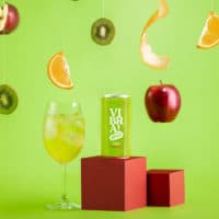 A foto apresenta o Vibra! Clericot da Evino em um fundo verde com um monte de frutas caindo e uma taça com o drink ao lado da lata.