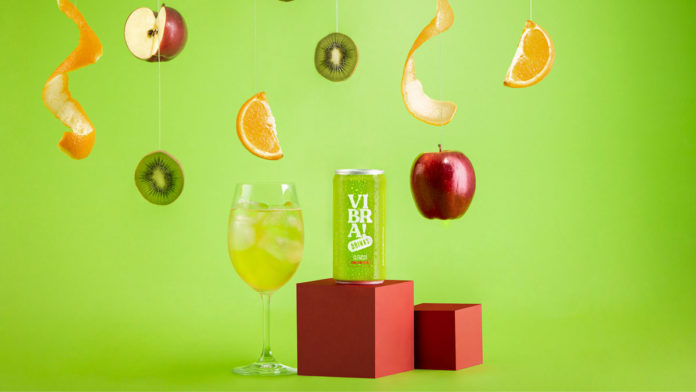 A foto apresenta o Vibra! Clericot da Evino em um fundo verde com um monte de frutas caindo e uma taça com o drink ao lado da lata.