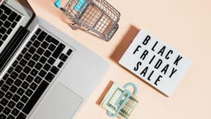 A foto apresenta uma mesa vista de cima com um notebook, uma placa escrito Black Friday Sale, um carrinho de compras e um dinheiro dobrado em um clipe de papel. Foto representativa para apresentar as extensões para uma Black Friday segura.