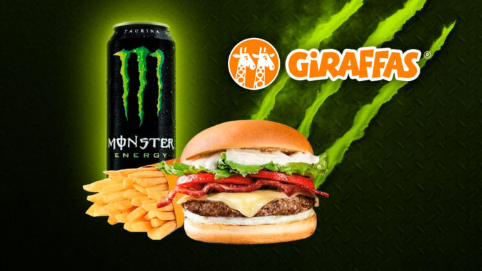 A foto apresenta o combo do Giraffas com o Moster Energy, com um fundo preto e os logos das marcas ao lado direito do sanduíche.