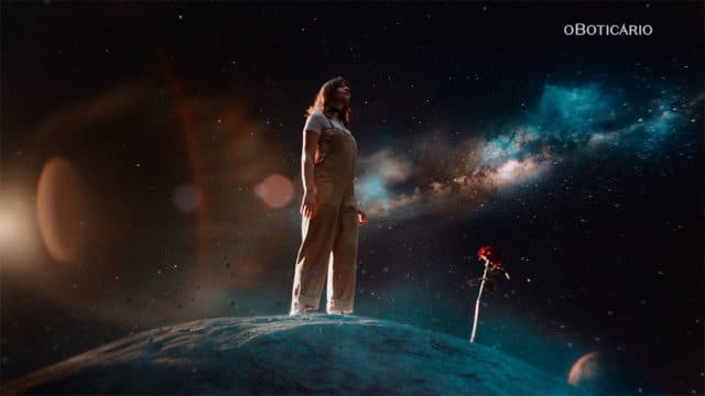 A foto apresenta um frame do filme da campanha sobre memória olfativa de O Boticário, a imagem apresenta uma mulher em outro mundo olhando para o horizonte ao lado de uma rosa.