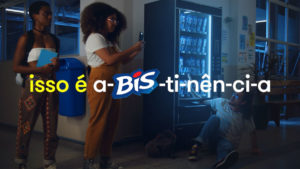A foto apresenta um dos filmes da campanha a-BIS-tinência de Bis, em que um homem está preso com o braço dentro de uma máquina de doces, enquanto duas garotas ficam olhando e tirando foto.