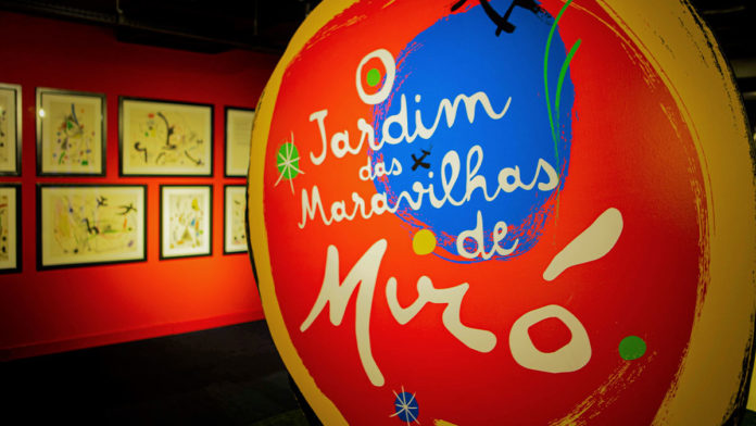 A foto apresenta o logo da exposição interativa de Joan Miró em um corredor com várias pinturas.