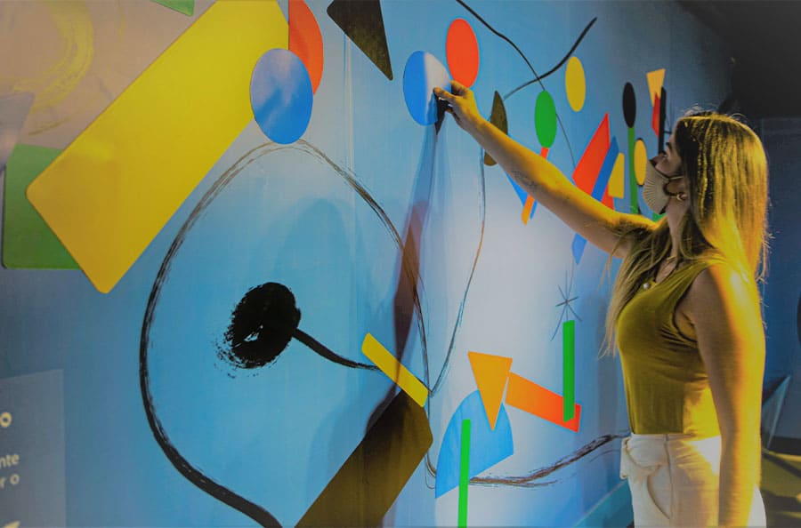 A foto apresenta uma mulher colando um adesivo na parede colorida.
