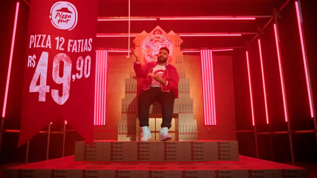 Frame da campanha com novo posicionamento da Pizza Hut. A foto apresenta Paulo Vieira sentado em um trono feito de caixas de pizza, ao lado de uma bandeira informando sobre a promoção.