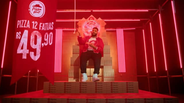 Frame da campanha com novo posicionamento da Pizza Hut. A foto apresenta Paulo Vieira sentado em um trono feito de caixas de pizza, ao lado de uma bandeira informando sobre a promoção.