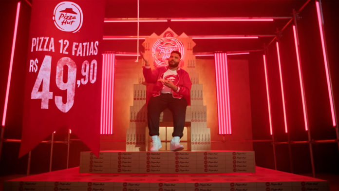 Frame da campanha com novo posicionamento da Pizza Hut. A foto apresenta Paulo Vieira sentado em um trono feito de caixas de pizza, ao lado de uma bandeira informando sobre a promoção.