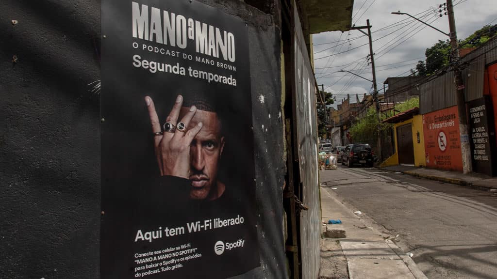 A foto apresenta um cartaz do Original Podcast Mano a Mano colado em uma parede dentro de uma periferia de SP.