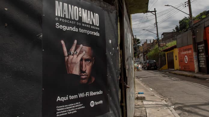 A foto apresenta um cartaz do Original Podcast Mano a Mano colado em uma parede dentro de uma periferia de SP.