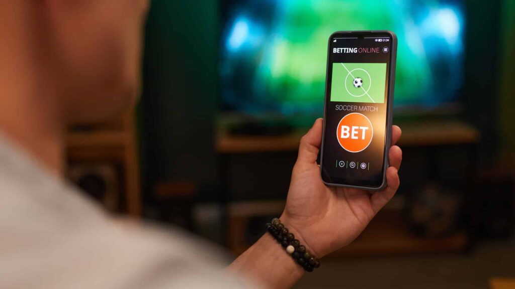 Aposta esportiva no celular com marca bet Pessoa segurando celular com aplicativo de aposta esportiva e marca de bet na tela em ambiente interno