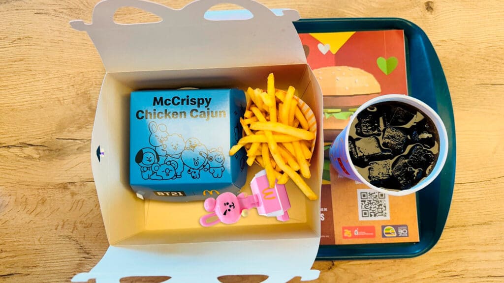 Imagem mostra em visão superior um combo do McDonald's trazendo caixa do McLanche Feliz, uma caixa do sanduíche, fritas, copo com refrigerante e bandeja. Ao fundo se vê uma mesa de madeira.