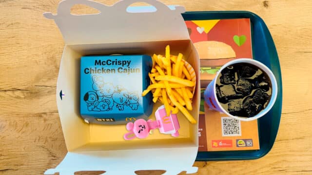 Imagem mostra em visão superior um combo do McDonald's trazendo caixa do McLanche Feliz, uma caixa do sanduíche, fritas, copo com refrigerante e bandeja. Ao fundo se vê uma mesa de madeira.