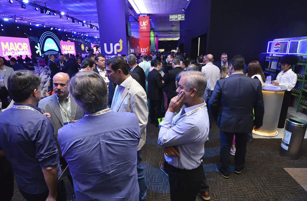 Multidão de profissionais interagindo em um evento de negócios, com telas de apresentação ao fundo e estandes de expositores.